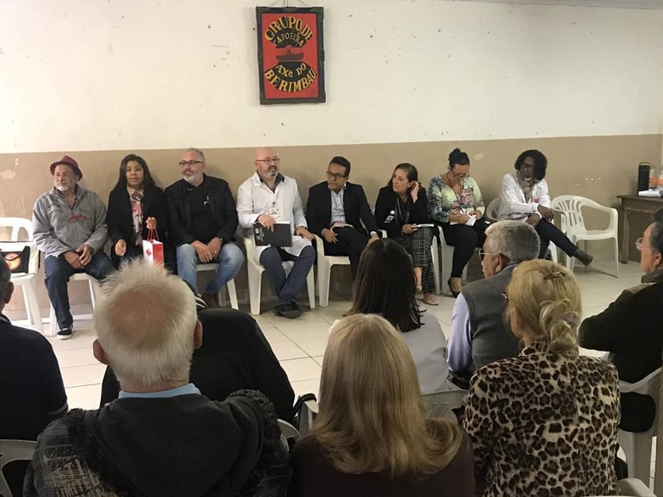 Mais de 16 pessoas estão sentados formando um “u”. O subprefeito, Gilmar Souza Santos é um deles. 
